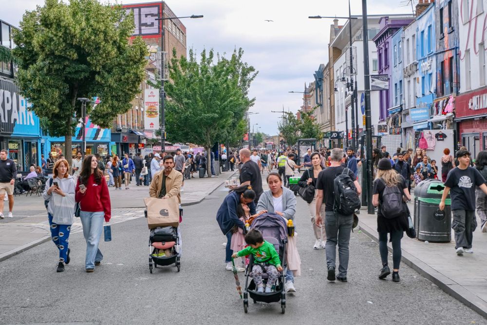 People walk, cycle, sit, and talk along a temporarily car-free stretch of Camden High Street in North London, which is lined with retail shopfronts, trees and flowers, terraces, bike parking, and benches.