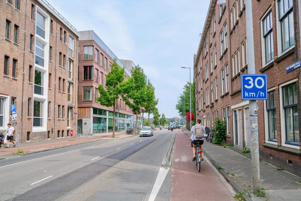 People of all ages and abilities walk, cycle, and drive along the low-traffic streets of Amsterdam, which are lined with buildings, trees, bike infrastructure, and prominent 30 km/hr signage.