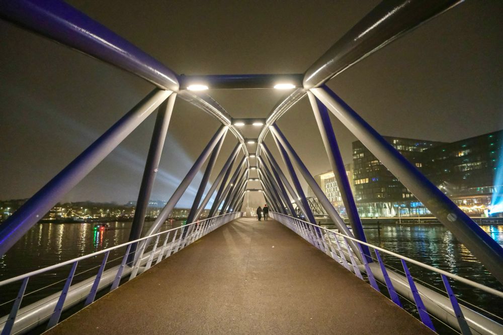 Pedestrians travel across a steel walking and cycling bridge in the harbor of Amsterdam on a dark and foggy night.