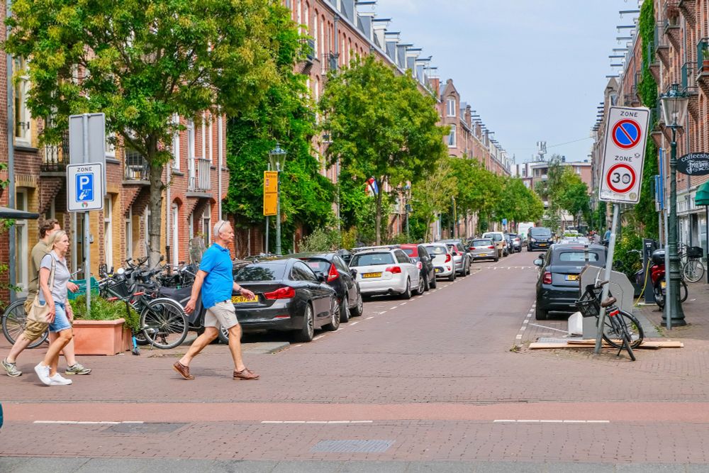 People of all ages and abilities walk, cycle, and drive along the low-traffic streets of Amsterdam, which are lined with buildings, trees, bike infrastructure, and prominent 30 km/hr signage.