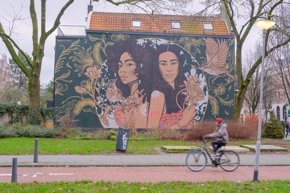 A cyclist rides along a red asphalt bike path through a park in Rotterdam. A large mural of two women is in the background.