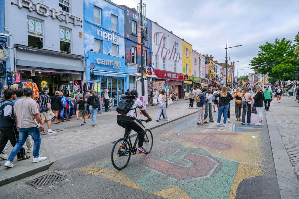 People walk, cycle, sit, and talk along a temporarily car-free stretch of Camden High Street in North London, which is lined with retail shopfronts, trees and flowers, terraces, bike parking, and benches.