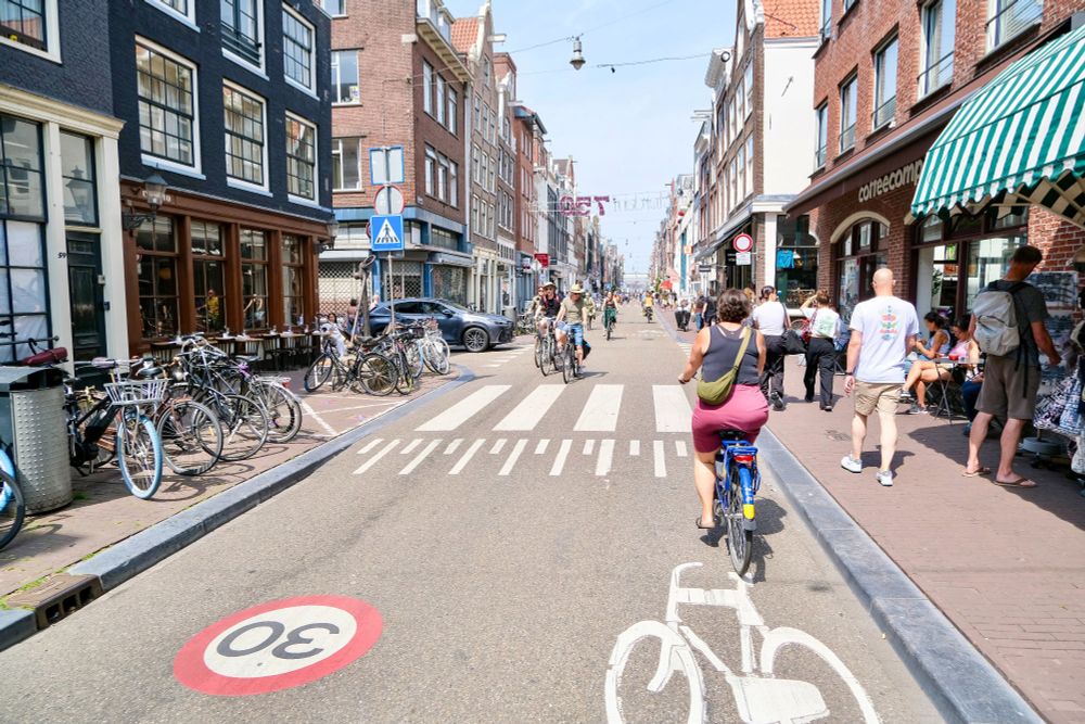 People of all ages and abilities walk, cycle, and drive along the low-traffic streets of Amsterdam, which are lined with buildings, trees, bike infrastructure, and prominent 30 km/hr signage.