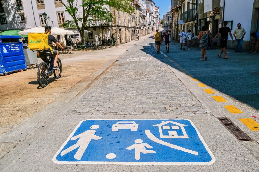 Crowds of people in casual summer attire walk and dine along the car-free streets lined with trees and terraces in the historic city centre of Santiago de Compostela.