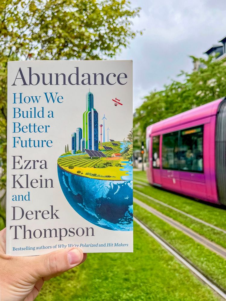 A hand holds a book, “Abundance: How We Build a Better Future” by Ezra Klein and Derek Thompson, while a pink tram passes over a grassy street in Reims, France out-of-focus in the background.