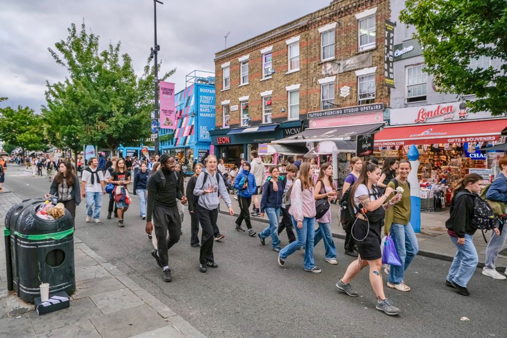 People walk, cycle, sit, and talk along a temporarily car-free stretch of Camden High Street in North London, which is lined with retail shopfronts, trees and flowers, terraces, bike parking, and benches.