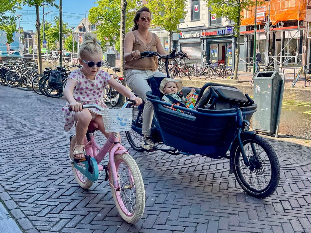 People of all ages and abilities in casual spring attire ride bicycles along dedicated infrastructure on the streets of Delft.