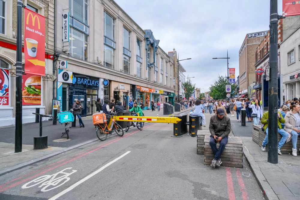 People walk, cycle, sit, and talk along a temporarily car-free stretch of Camden High Street in North London, which is lined with retail shopfronts, trees and flowers, terraces, bike parking, and benches.