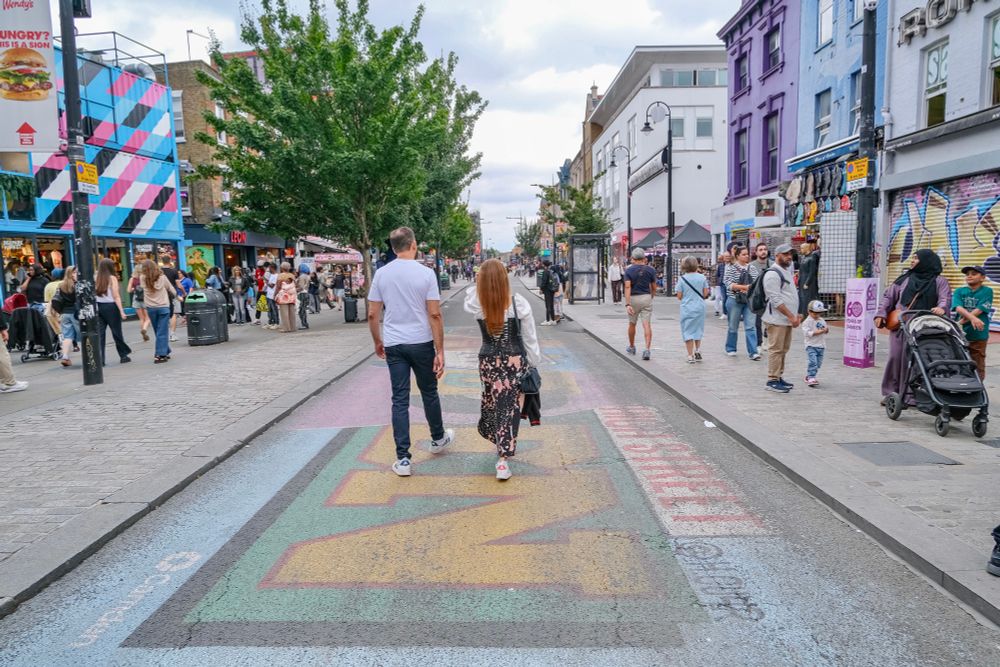 People walk, cycle, sit, and talk along a temporarily car-free stretch of Camden High Street in North London, which is lined with retail shopfronts, trees and flowers, terraces, bike parking, and benches.