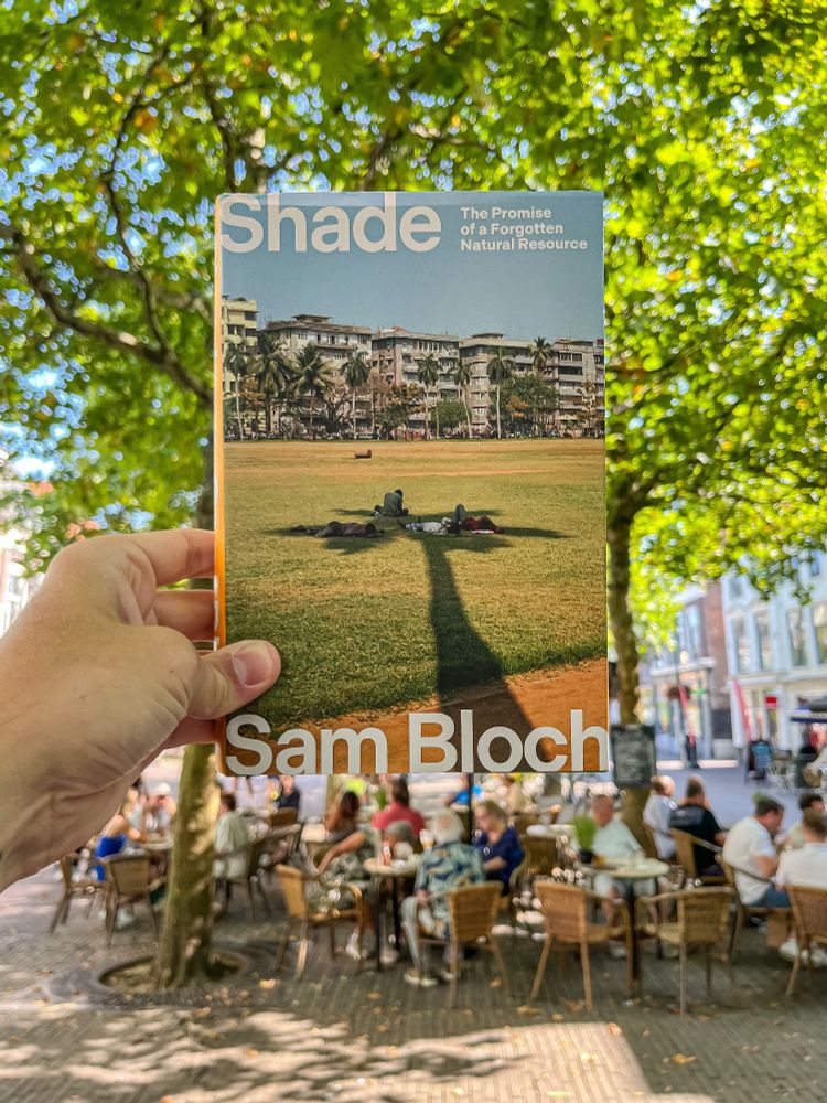 A hand holds a book, “Shade: The Promise of a Forgotten Natural Resource” by Sam Bloch, with a busy terrace below a canopy of street trees out-of-focus in the background.  