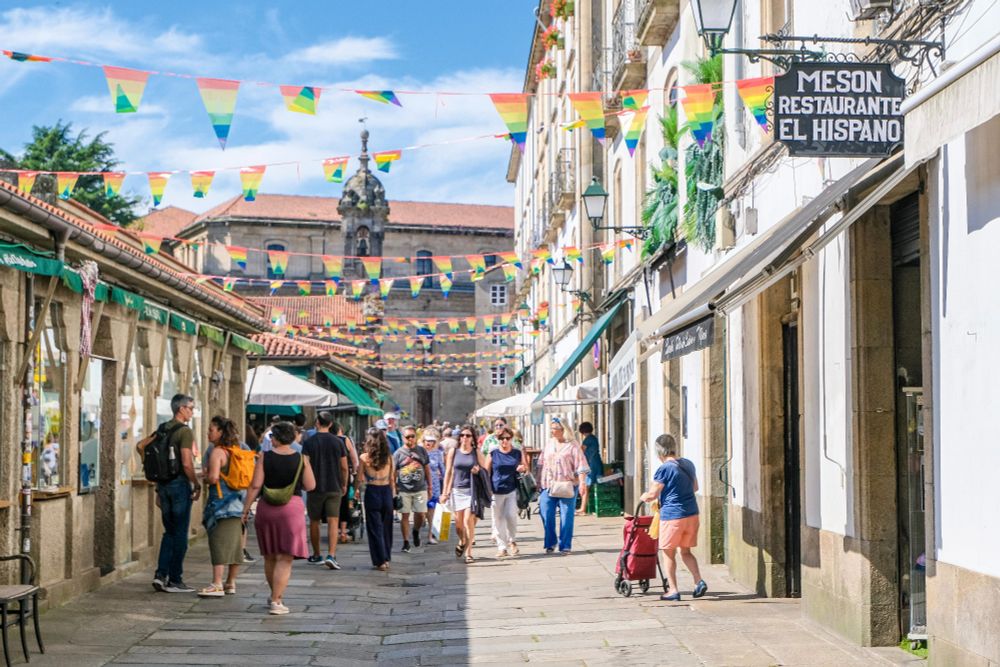 Crowds of people in casual summer attire walk and dine along the car-free streets lined with trees and terraces in the historic city centre of Santiago de Compostela.