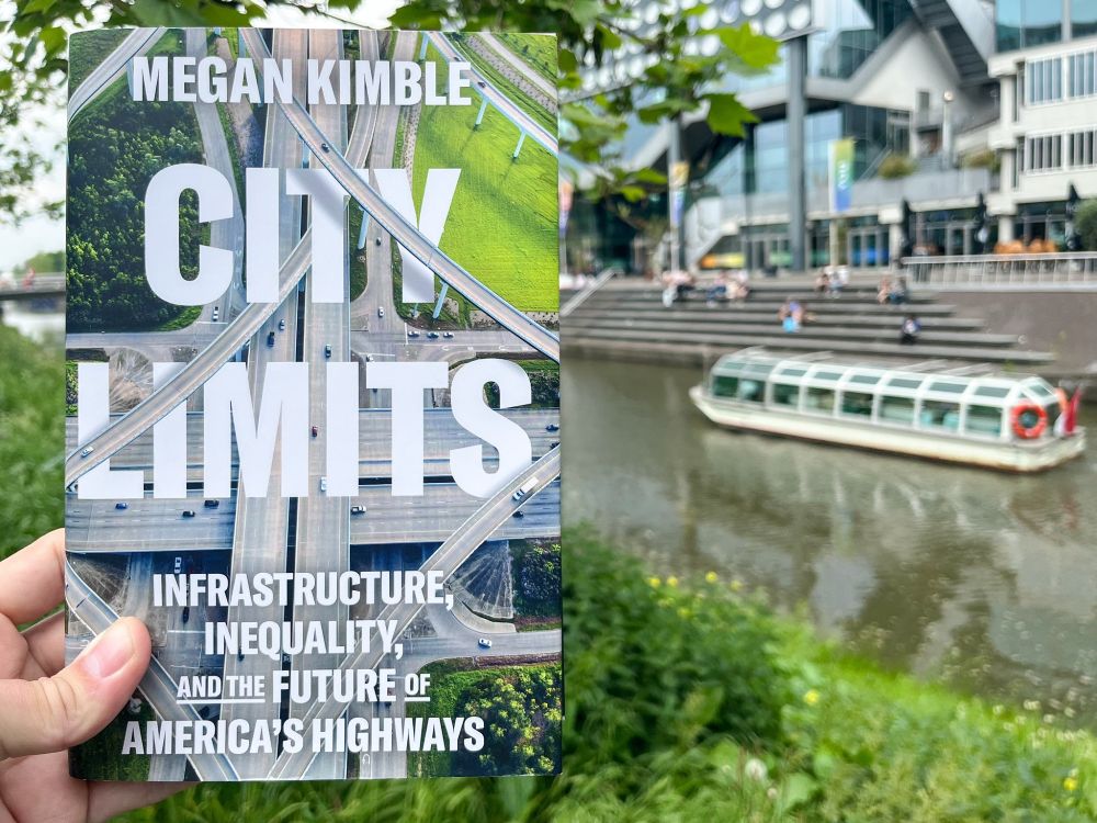 A photograph of a hand holding a book about city planning in front of an urban streetscape.