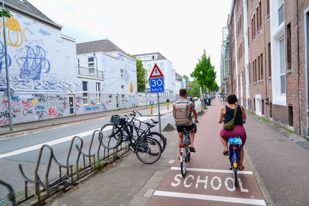 People of all ages and abilities walk, cycle, and drive along the low-traffic streets of Amsterdam, which are lined with buildings, trees, bike infrastructure, and prominent 30 km/hr signage.