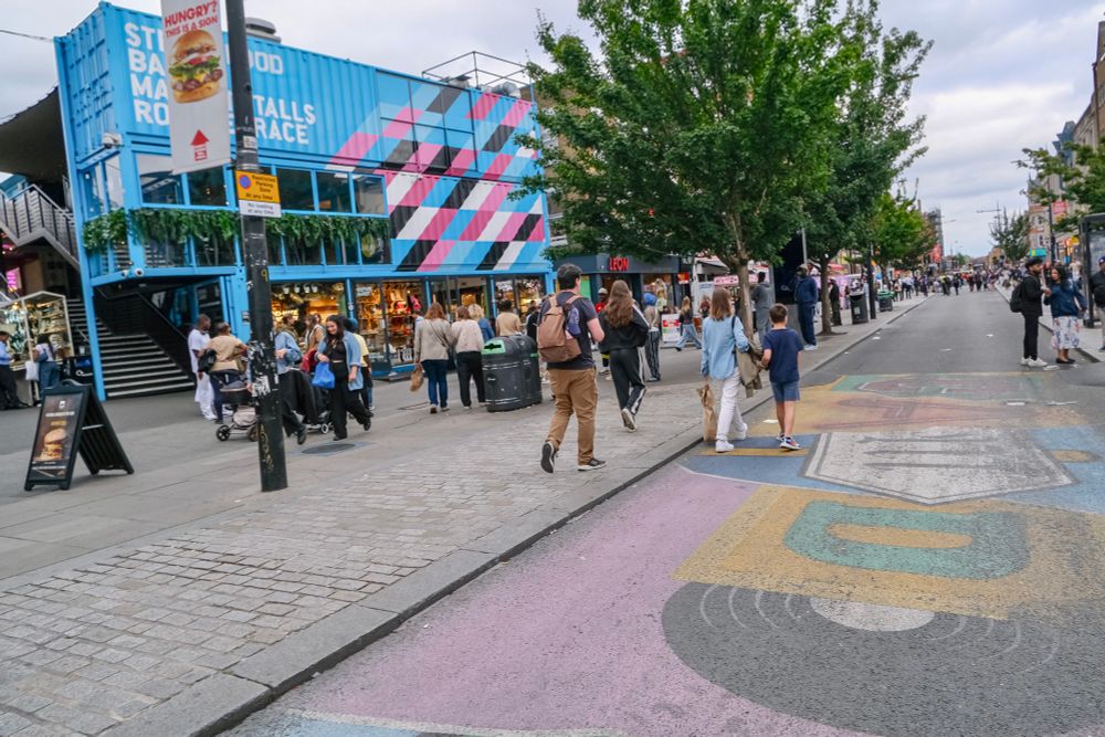People walk, cycle, sit, and talk along a temporarily car-free stretch of Camden High Street in North London, which is lined with retail shopfronts, trees and flowers, terraces, bike parking, and benches.