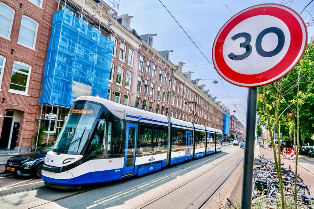 People of all ages and abilities walk, cycle, and drive along the low-traffic streets of Amsterdam, which are lined with buildings, trees, bike infrastructure, and prominent 30 km/hr signage.