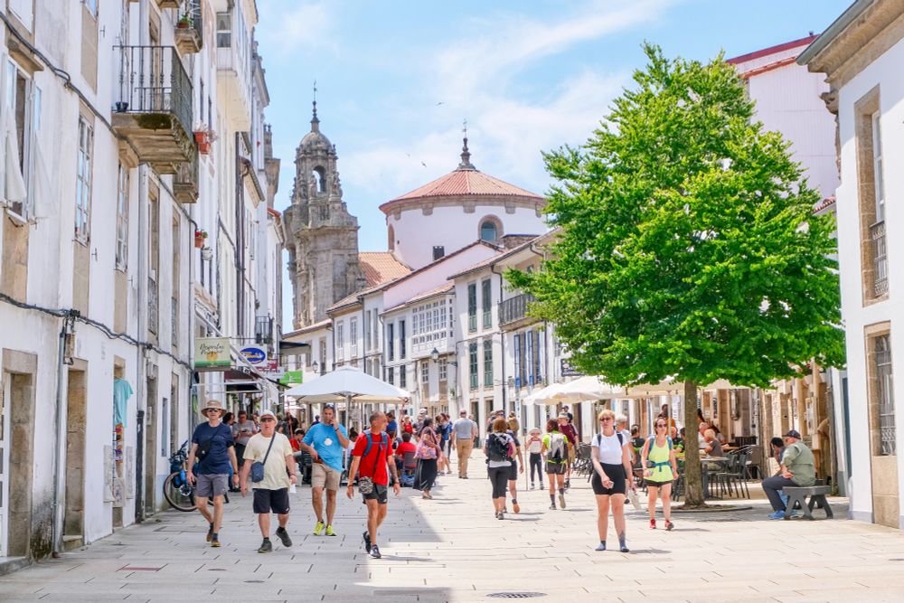 Crowds of people in casual summer attire walk and dine along the car-free streets lined with trees and terraces in the historic city centre of Santiago de Compostela.