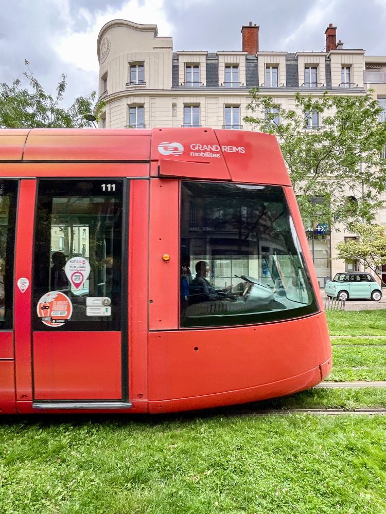 Various photos of colourful trams moving along grassy streets lined with trees and vegetation in Reims, France.