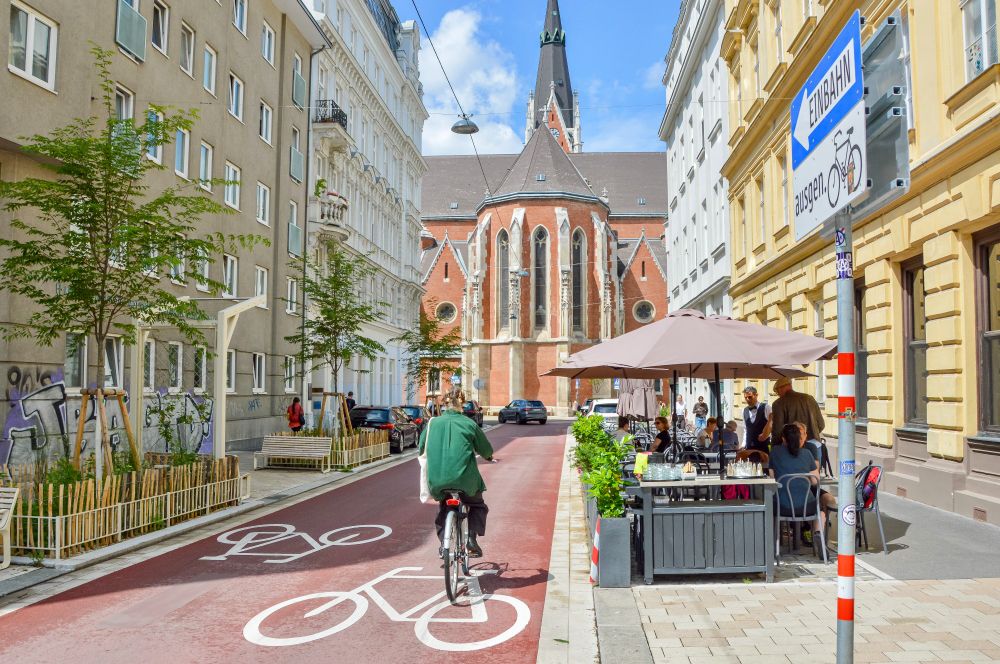 People in casual spring attire cycle on Argentinierstraße in Vienna—a red asphalt “bicycle street” lined with four-storey buildings, trees and plants, and wooden benches. 