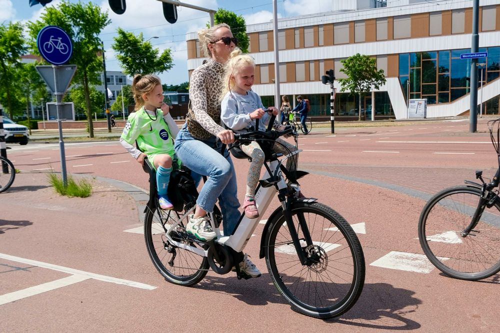 A woman rides a bicycle with a young child sitting on both the front and rear rack along a protected bike lane in Delft.