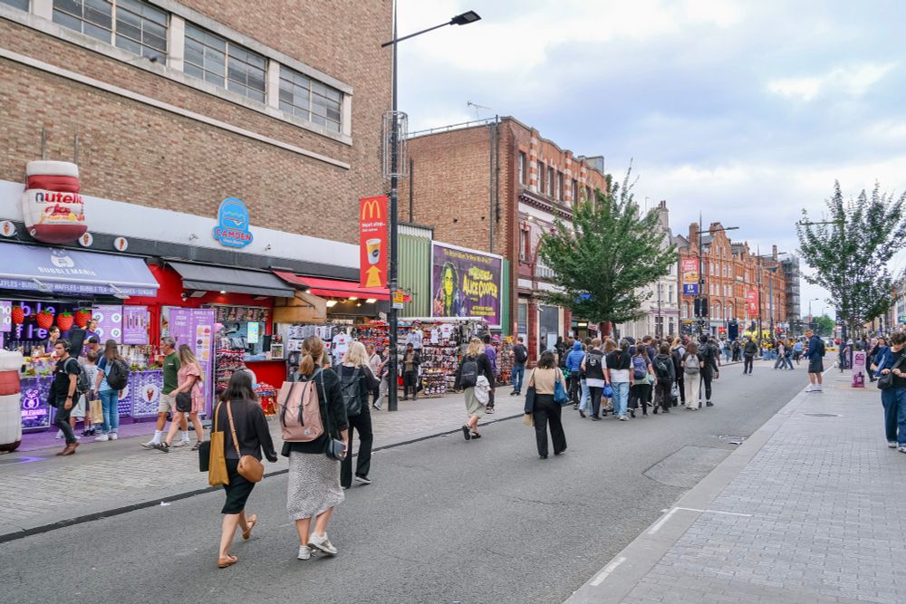People walk, cycle, sit, and talk along a temporarily car-free stretch of Camden High Street in North London, which is lined with retail shopfronts, trees and flowers, terraces, bike parking, and benches.