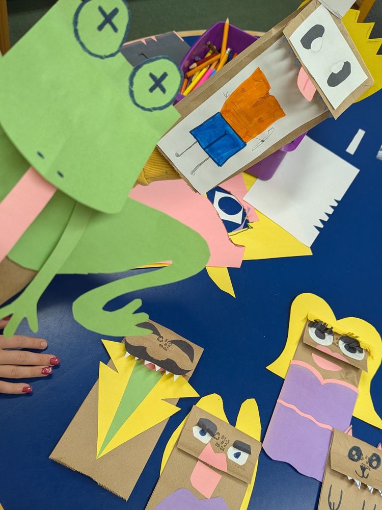 Photograph of puppets made with small brown paper bags and coloured craft paper, featuring a frog, Bart Simpson, a werewolf in a suit, 2 Dolly Partons, and a cat. 