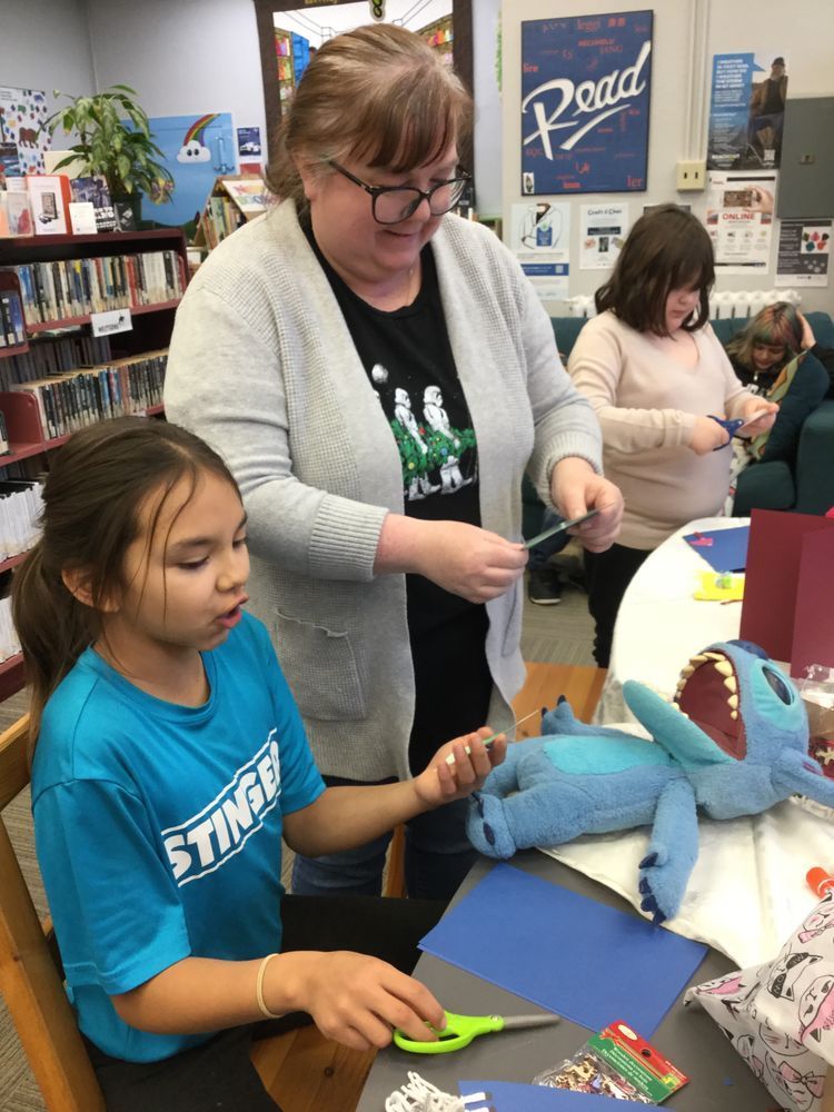 a woman standing and smiling, she is wearing a grey cosy sweater. she is showing 2 young girls how to make a paper craft