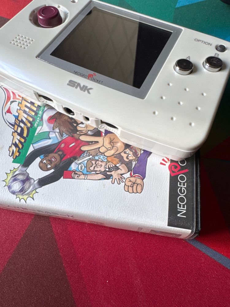 Photo of a NeoGeo Pocket, a retro handheld gaming device by the company SNK. It is unpacked, turned off and sitting on top off the game case of the game NeoGeo Cup ’98, s soccer game for the device. 