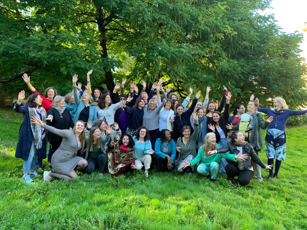 Een groep WECF medewerkers die vrolijk lachen en hun handen in de lucht houden bij het poseren voor de foto. De mensen zitten en staan in twee rijen op een grasveld met bomen in de achtergrond. 