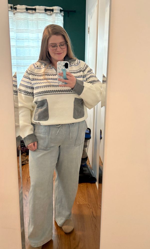 A mirror selfie of me, a white woman with long blonde hair, wearing light wash wide leg jeans, tan clogs, and the folklore sweater from Taylor Swifts holiday merch collection. 