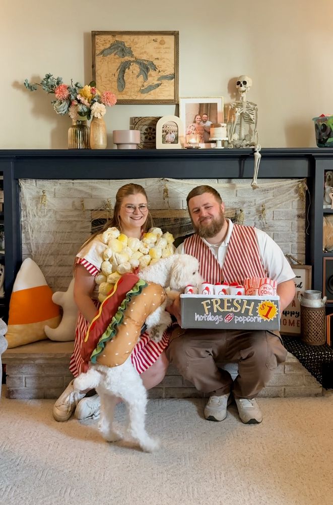 Me, my husband, and my dog sitting on a fireplace hearth while dressed up for halloween. I am a bag of popcorn, my dog is a hotdog, and my husband is a vendor selling hotdogs and popcorn for $1