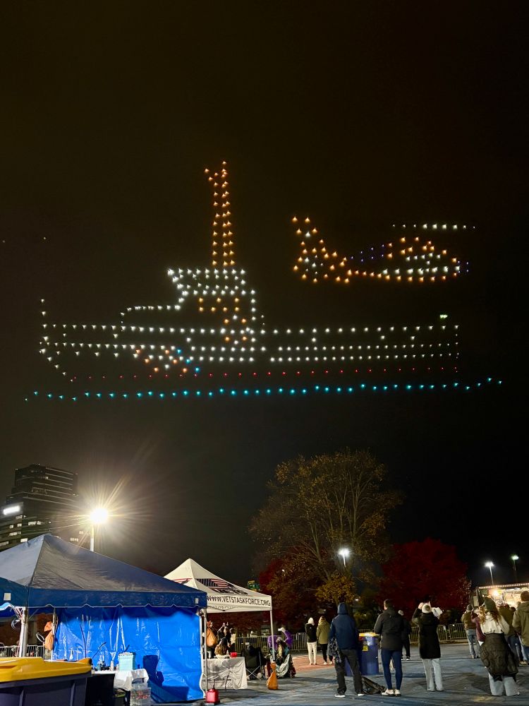 500 drones forming an image of an aircraft carrier ship with a helicopter taking off from it over a small gathering of people