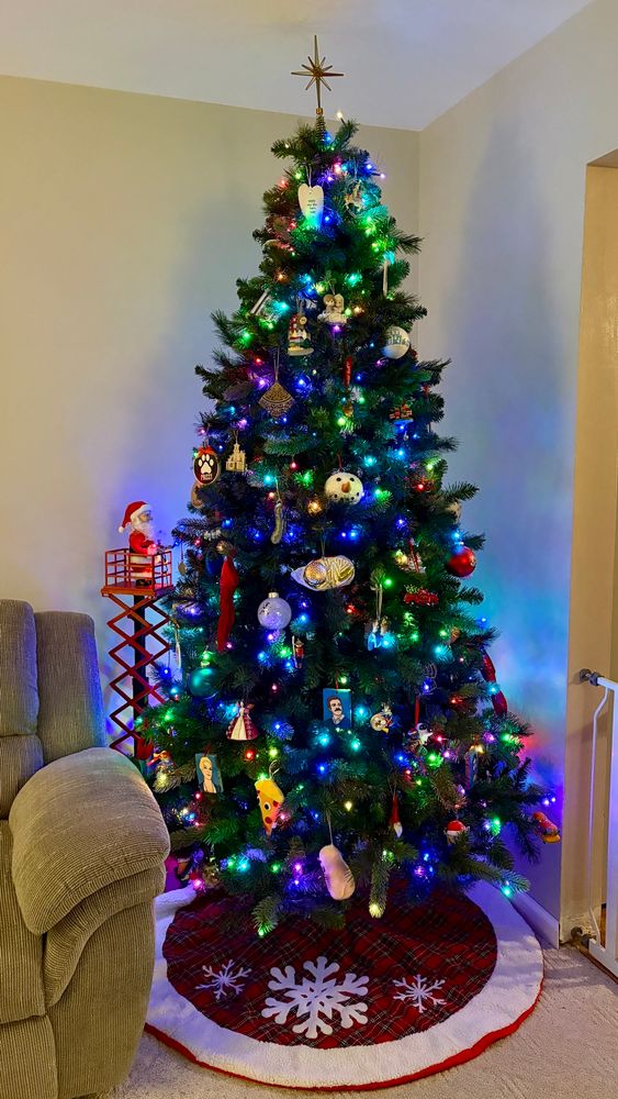 A Christmas tree glowing with multi colored twinkling lights, covered in a random smorgasbord of ornaments. There is a Santa on a scissor lift hanging lights behind the tree, and there is a plaid tree skirt adorned with snowflakes beneath 