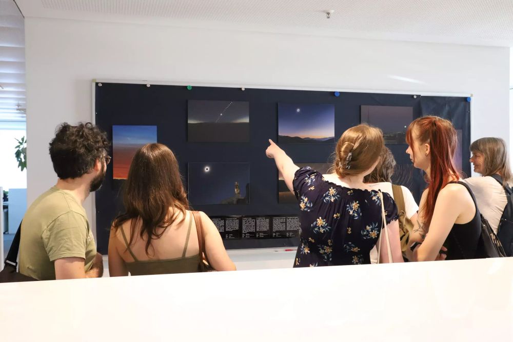 Ein Whiteboard an dem Fotografien hängen. Die Bilder zeigen verschiedene Weitwinkelaufnahmen einer Sonnenfinsternis. Vor der Tafel, die Rücken dem Fotografen zugewandt, stehen mehrere Menschen, betrachten die Bilder und zeigen darauf.