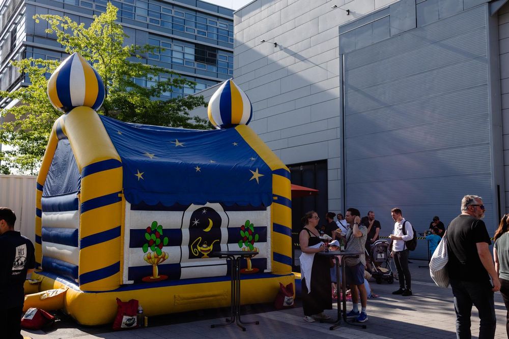 Fill view of a blue-and-yellow bouncy castle in front of a large white building in the background. People are swarming around it.