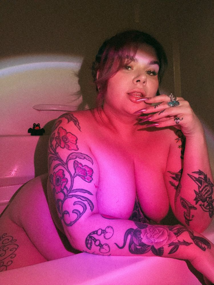 Bratty in the bath with pink lighting touching her nails to her lips 