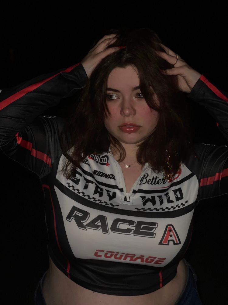 A picture of me outside at night with the flash on. I have tinsel in my hair and am wearing a cropped quarter zip that looks inspired by a race car driver. My hands are in my hair and I’m looking off to the side