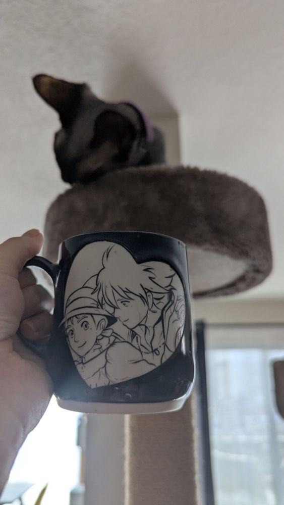 I'm holding an 8 oz navy blue handmade ceramic mug in the air. The mug features a hand painted Sophie and Howl from Howls Moving Castle within a heart. Howl is leaning over Sophie's shoulder, which is a scene from the beginning of the movie where he saves her from some military men and they fly above the city while holding hands. Behind the mug is my Sphynx cat Zara's cat tree and she's rubbing her face on her tree. 