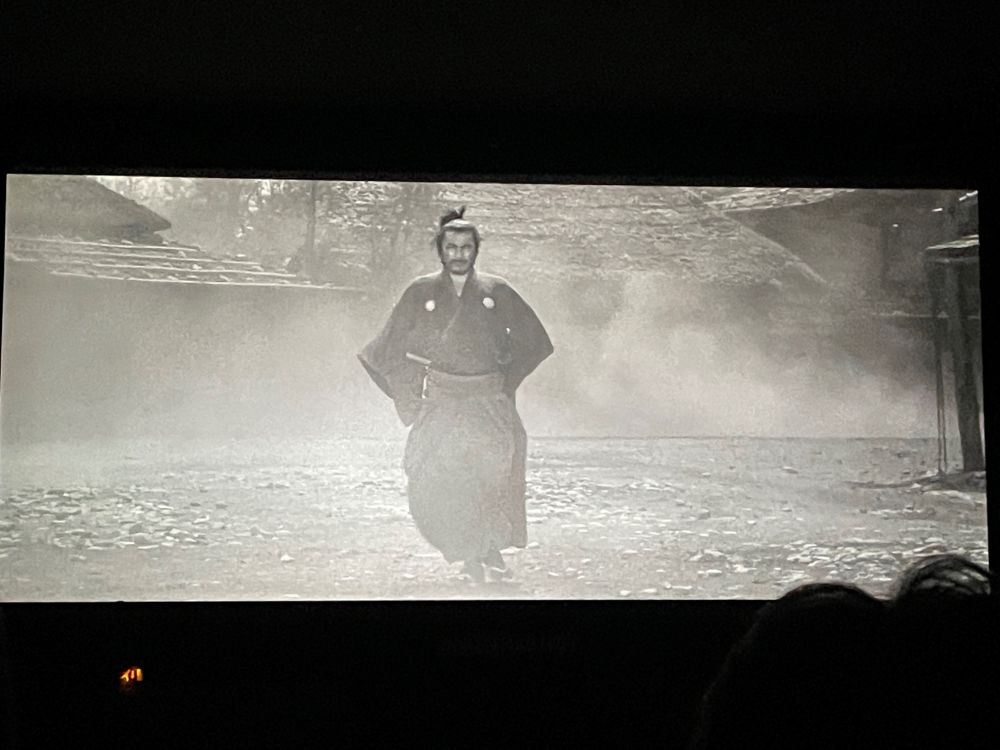 A scene from Yojimbo. The lead character who plays a samurai stands in the middle of a desolate street with dust billowing around him.
