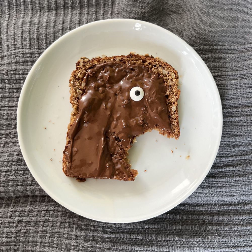 Angebissenes Brot mit Schokocreme. Durch den Biss rechts unten und ein Zuckerauge auf dem Brot entsteht der Eindruck eines schreienden Gesichts. 