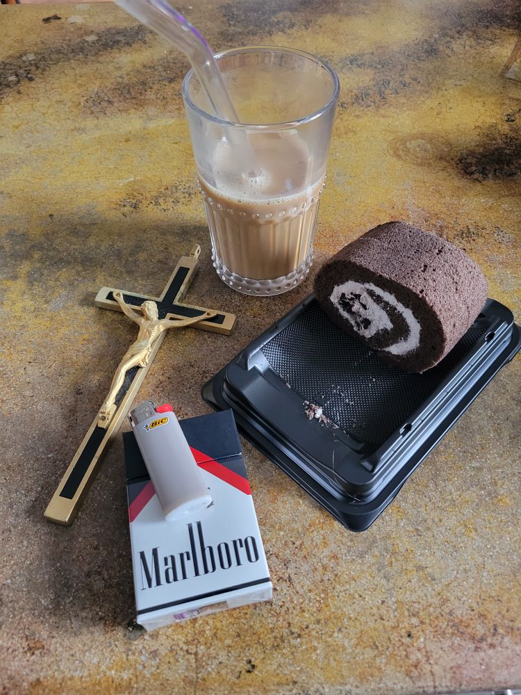 a swiss roll cake, a pack of Marlboro black cigarettes and lighter, a half full glass of iced coffee, and a crucifix on a weird looking yellow kitchen counter