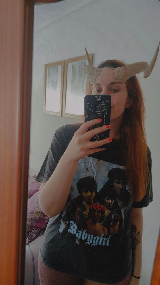 A photo of me in front of a mirror, wearing a T-shirt with several in-game images of Gortash (Baldur's Gate 3) and the words 'BABYGIRL' on it, and with bone white tiefling horns.