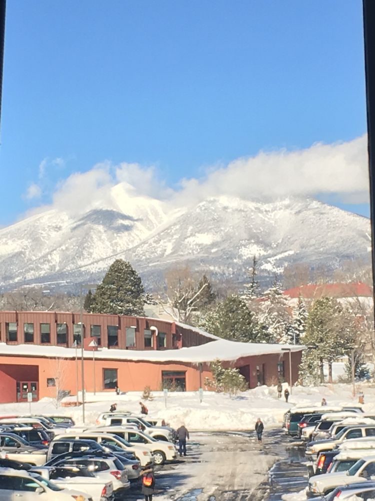 Snow-covered San Francisco peaks. In the foreground is a parking lot and building. 