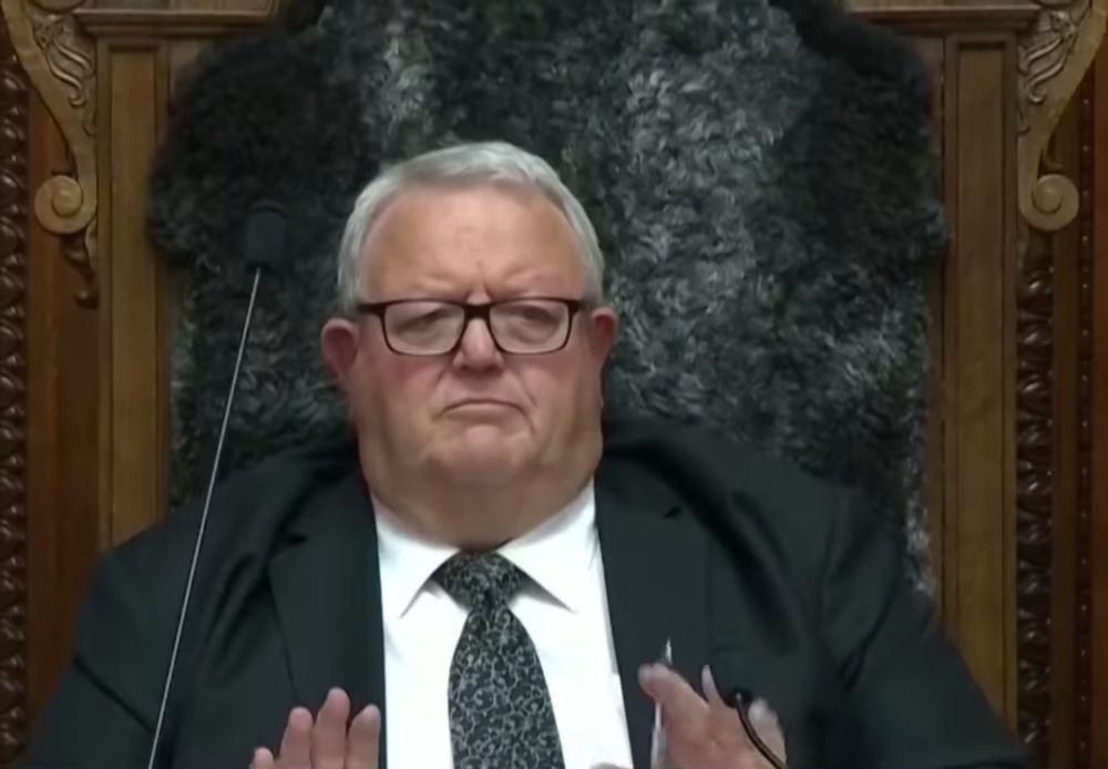 A close-up of the Speaker of the New Zealand Parliament seated in a formal chair lined with fur. He is a pale obese man, his neck overflowing the collar of his expensive but poorly fitted suit. He has his little hands held in front of him as if he wants to push this situation away from him as he can. He’s rolling his eyes, revealing irritation, condescension, and frustration as he observes the events unfolding in the chamber.