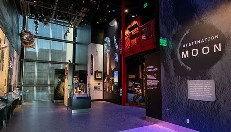 View of the "Destination Moon" exhibit at the Smithsonian National Air and Space Museum. The exhibit features dramatic lighting, large wall graphics of astronauts and rockets, and interpretive panels explaining the Apollo missions. Artifacts and models are displayed in cases along the walkway, capturing the history and ambition of the space race.