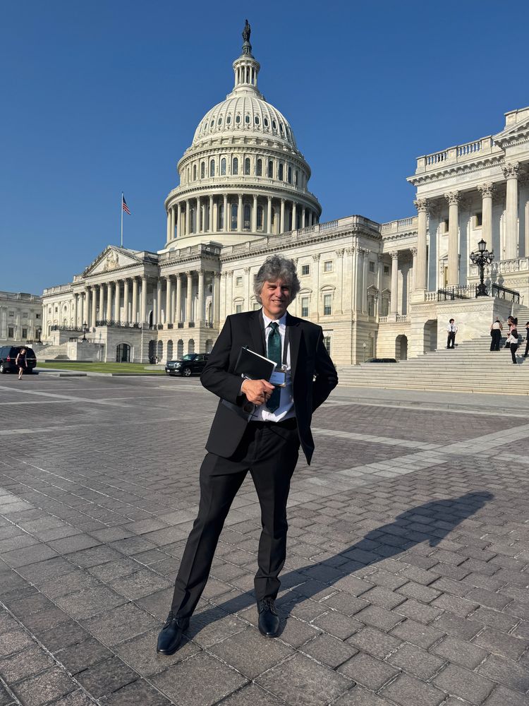 A man in a black suit stands confidently in front of the U.S. Capitol building on a sunny day, holding a folder and wearing a name badge. The iconic dome rises behind him against a clear blue sky. The brick plaza in the foreground casts a long shadow, adding depth to the image.