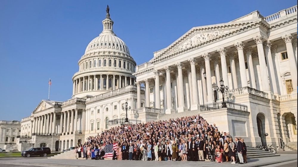 Hundreds of Citizens’ Climate Lobby volunteers gather on the steps of the U.S. Capitol for a group photo during lobby day. The iconic dome rises prominently in the background under a clear blue sky, while participants in business attire stand shoulder to shoulder, holding banners and American flags to mark the occasion.