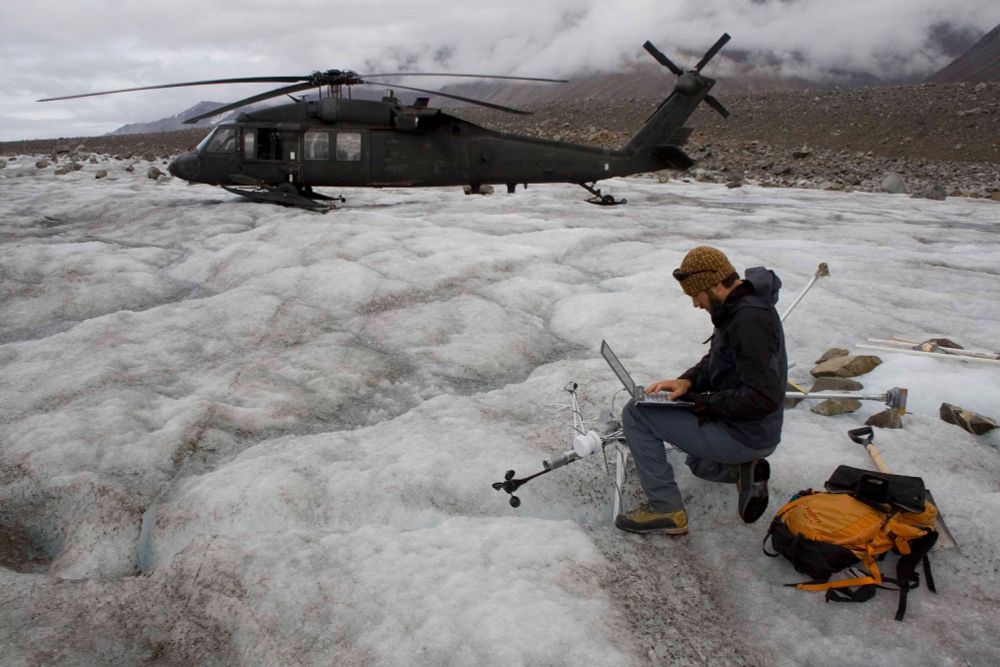 A man kneeling on a glacier and working on a laptop next to a stupid, fallen-over weather station, and with a US Army Blackhawk helicopter in the background.