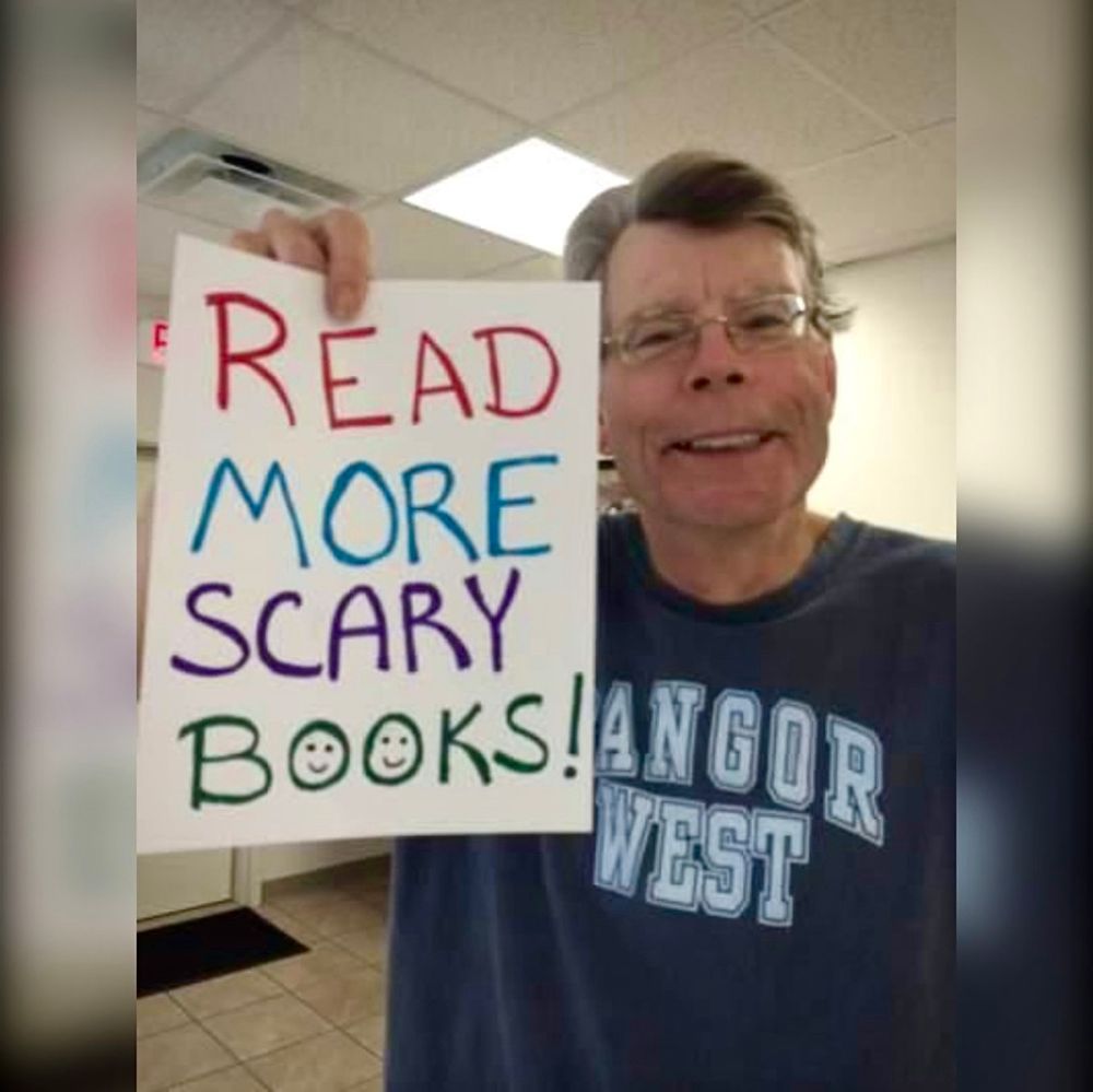 Stephen King holding a hand-written sign that says “Read more scary books!”