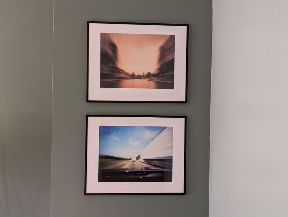 Two framed photographs hang vertically on a dark green wall, near the corner where a light green wall begins on the right. The top image features an abstract, motion-blurred cityscape in warm tones. The bottom image shows a motion-blurred highway scene in cooler tones, with a road and a white truck visible in the distance.