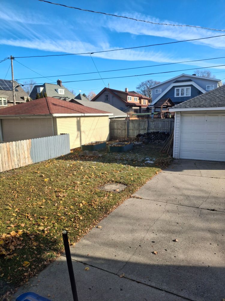 View of a narrow urban neighborhood back yard. It is a sunny day, the fall lawn is sprinkled here and there with yellow maple leaves. To the right is a concrete driveway leading to a small garage. In the back of the yard are two green metal raised gardening containers. A neighbor's garage abuts this gardening area. Over the tops of the garages we see the roofs of several neighboring houses.